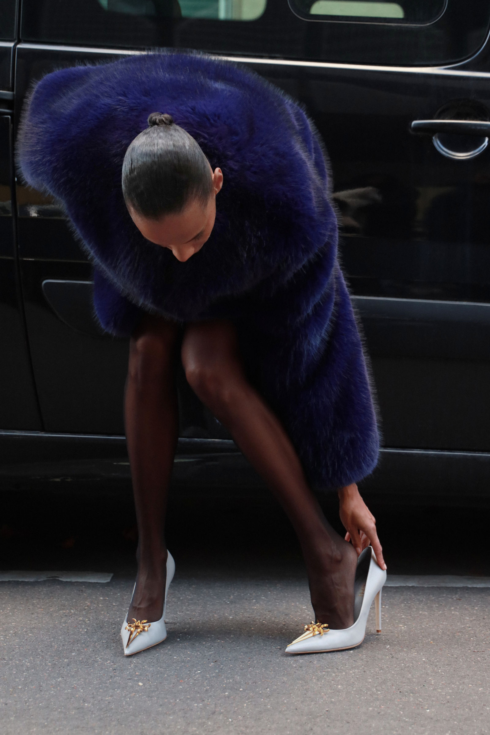 Person wearing a blue fur coat and high heels in front of a black vehicle